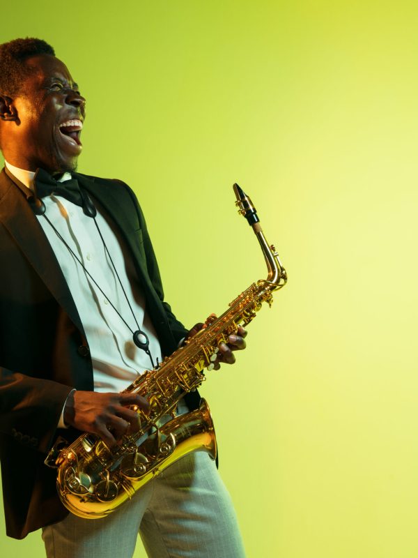 Young african-american jazz musician playing the saxophone on gradient yellow-green studio background. Concept of music, hobby, festival. Joyful attractive guy improvising. Colorful portrait of artist.