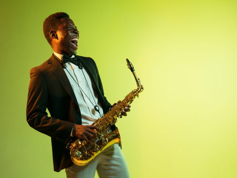 Young african-american jazz musician playing the saxophone on gradient yellow-green studio background. Concept of music, hobby, festival. Joyful attractive guy improvising. Colorful portrait of artist.