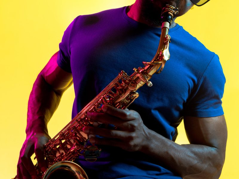 Young african-american jazz musician playing the saxophone on yellow studio background in trendy neon light. Concept of music, hobby. Joyful attractive guy improvising. Colorful portrait of artist.