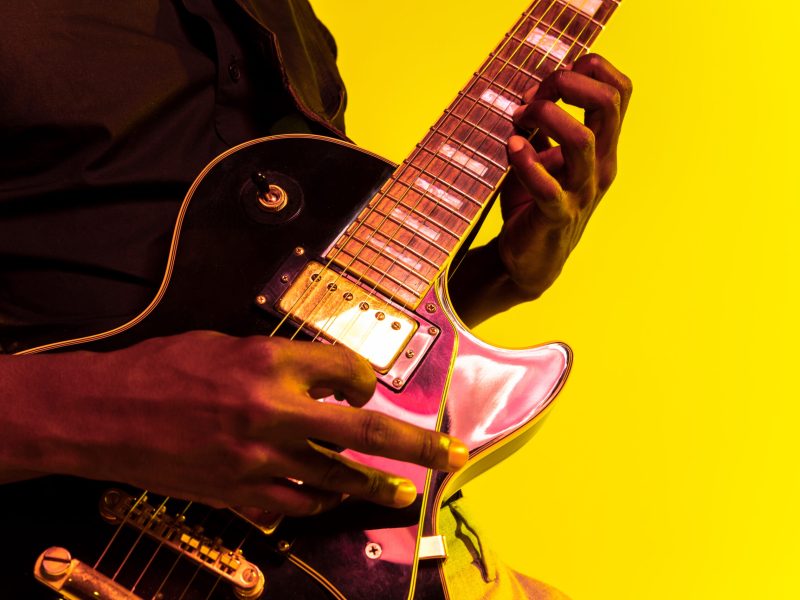 Young african-american musician playing the guitar like a rockstar on yellow background in neon light. Concept of music, hobby, festival, open-air. Joyful attractive guy improvising, singing song.